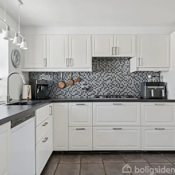 Hvidt køkken med sorte bordplader. Kaffemaskine og brødrister står på bordet. Mosaikfliser pryder væggen over komfuret. Store vinduer giver lys, og lamper hænger ovenfor.