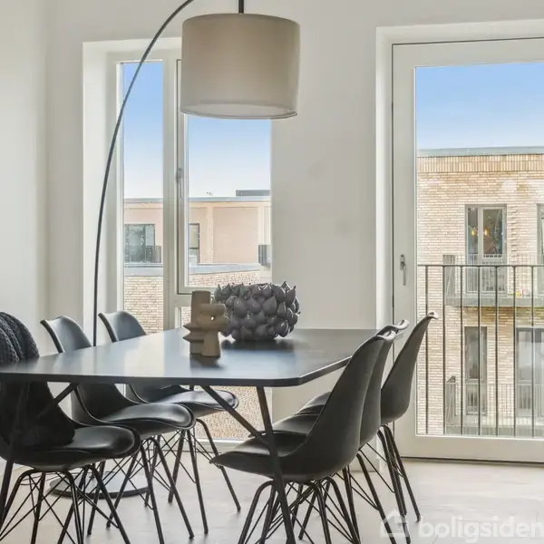 Et sort spisebord står i en lys spisestue med seks stole omkring. En moderne lampe hænger over bordet, og der ses udsigt til en bygning gennem glasdøren og vinduet.