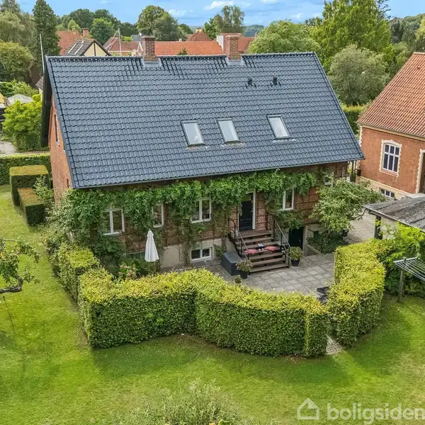 Et stort murstenshus med skråt tag dækket af tagvinduer, omkranset af en velplejet have med hække og træer. En terrasse med havemøbler foran huset.