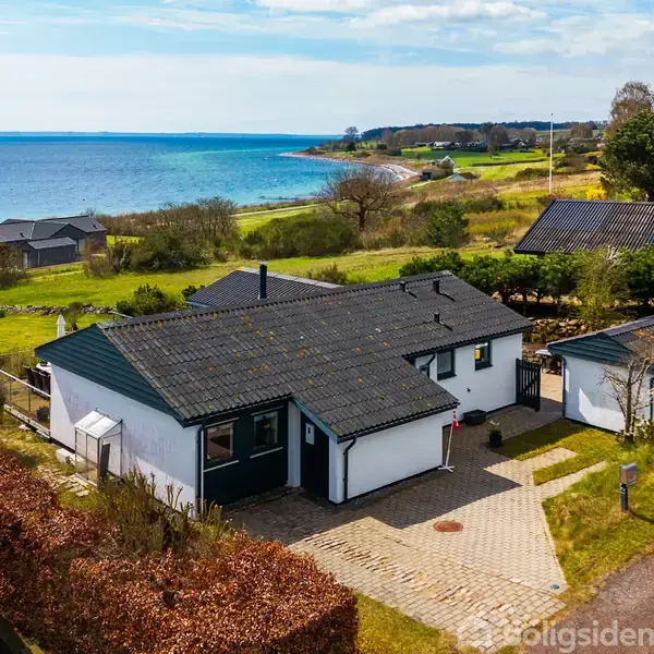 Hus i hvidt med sort tag står på en stenbelagt indkørsel ved havet. Grønt landskab og kystlinje ses i baggrunden under en blå himmel med lette skyer.