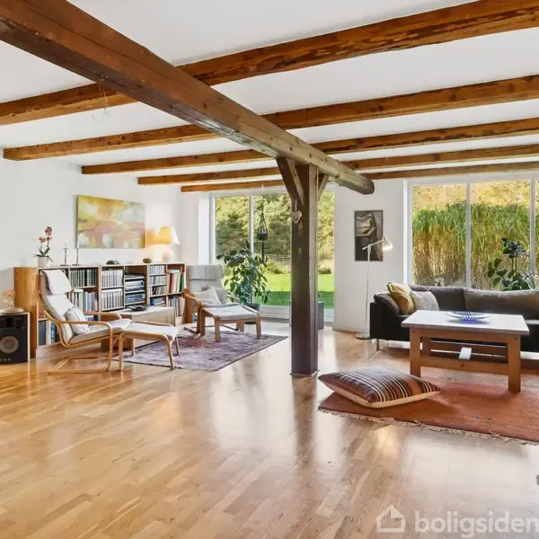 Stue med trægulv og synlige loftsbjælker. To stole og en reol står mod en glasdør til haven. En hjørnesofa med sofabord er placeret ved store vinduer med udsigt til grønne omgivelser.