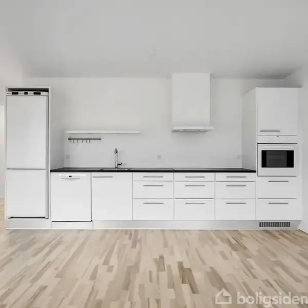 Køkken med hvide skabe, indbygget ovn og emhætte, placeret mod en hvid væg. Træplankegulv skaber minimalistisk og lys atmosfære. En åben dør fører til et tilstødende rum.