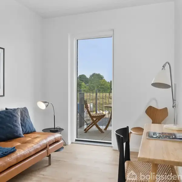 Et lyst rum har en brun lædersofa med blå puder, en gulvlampe, et skrivebord med stol og lampe. En altandør med udsigt til grønne træer åbner til en lille balkon.