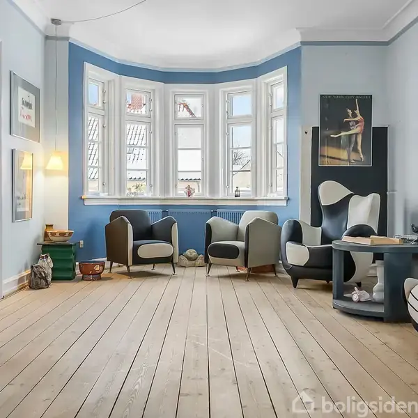 En stue med lyseblå vægge indeholder moderne møbler, herunder lænestole og et sofabord, placeret ved et stort karnapvindue. Trægulvet giver rummet en varm atmosfære.