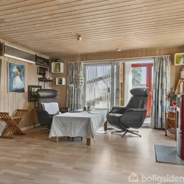 Bekvemme lænestole står ved et bord med dug i en stue med trævægge og loft. Væggene er dekoreret med malerier, og der er glasdøre, der slipper lys ind.