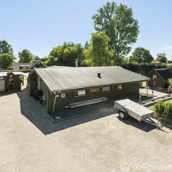 Et grønt sommerhus står på en brolagt gårdsplads, omgivet af græs og hække. En trailer er parkeret ved siden af huset. Baggrunden viser flerårige træer og klart blå himmel.