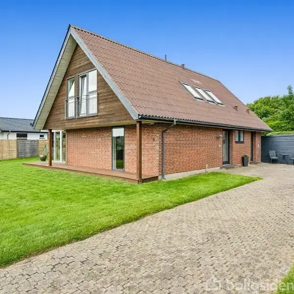 Et rødt murstenshus med brunt saddeltag står i en velholdt have med græsplæne, flisebelagt indkørsel og træhegn. Flere tagvinduer ses, og der er en terrasse foran huset.