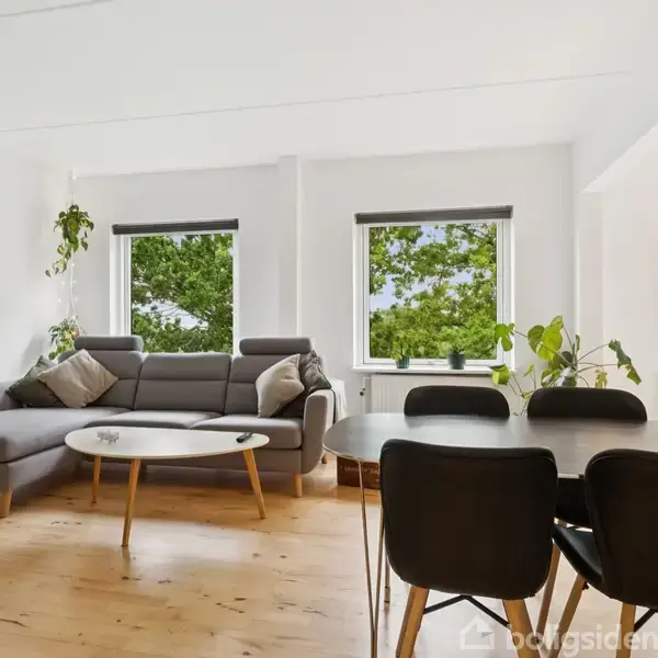 En grå sofa står i en lys stue ved siden af et rundt sofabord. Vinduerne viser grønne trætoppe, mens spisebordsstole er placeret omkring et bord til højre.