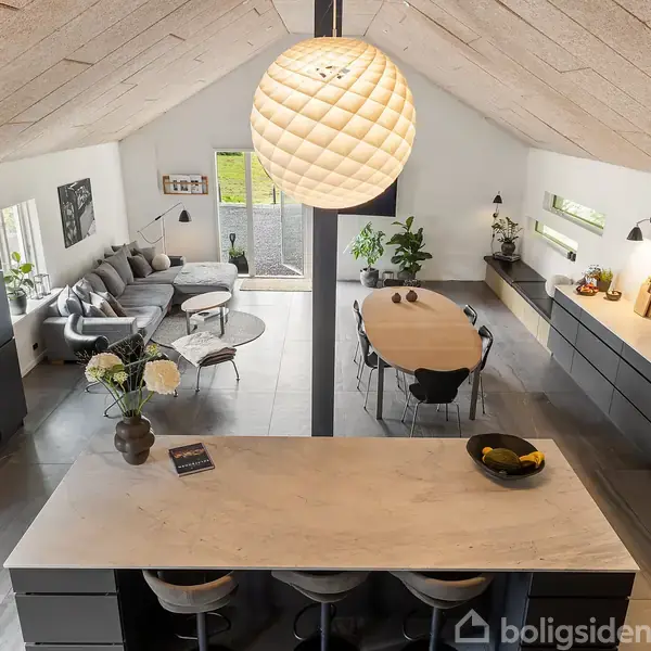 En moderne åben stue og køkken med spisebord, tre barstole ved køkkenøen, grå sofa og sofabord mod venstre. Store vinduer giver naturligt lys, indrettet med planter.