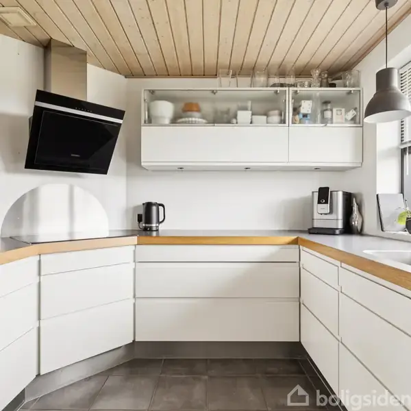 Hvidt køkken med L-formet bordplade og indbygget vask. Der står en elkedel og kaffemaskine på bordet. Ovenfor er der vægskabe, og et stort vindue giver naturligt lys.