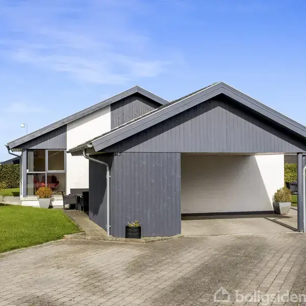 Et moderne hus med grå facader har en indkørsel og carport i forgrunden. Haven omkring er grønt anlagt med små træer, under en klar blå himmel.