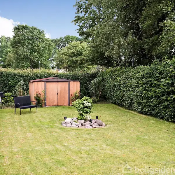En velplejet græsplæne med et lille bålsted i midten. En træbænk og grill står tæt ved et skur omgivet af tætte, grønne hække og træer.