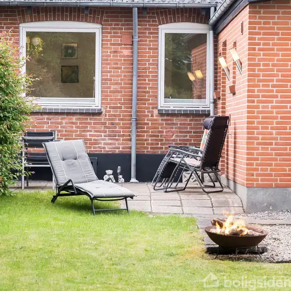 En liggestol står på en grøn græsplæne foran et rødt murstenshus med store vinduer. Ved siden af er der en brændende udendørs ildskål på grus.