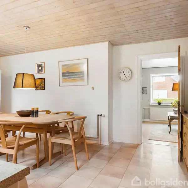 Et spisebord med stole står i et køkken med lyst træloft. Et køleskab er i hjørnet. På væggene hænger billeder og en rund ur. En kommode står ved siden af en døråbning.