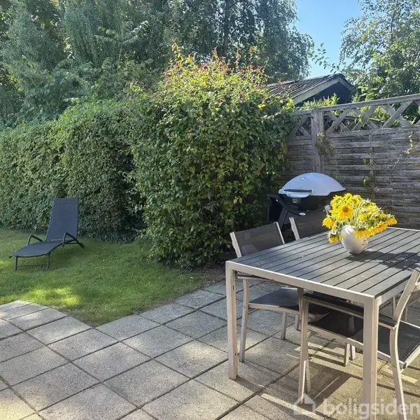 En terrasse har et bord med stole og en vase med gule blomster. Ved siden af står en grill. En liggestol er placeret på græsset ved en hæk.