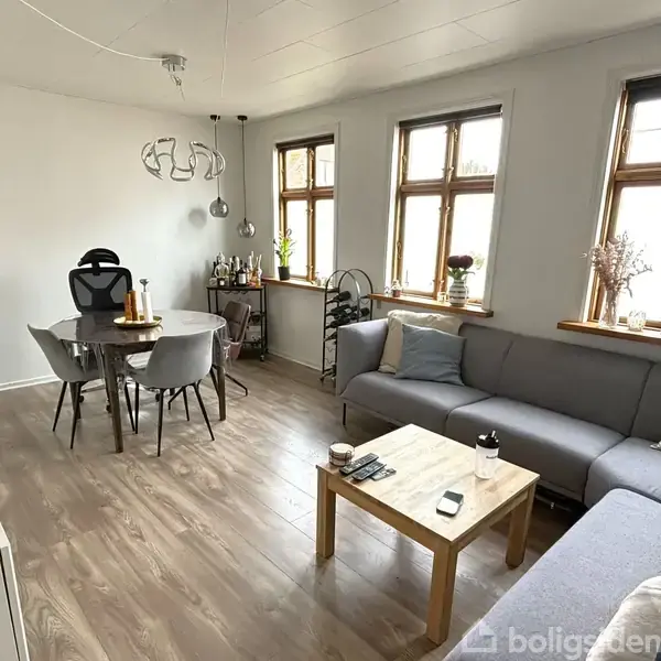 Stue med gråt hjørnesofaarrangement rundt et trækaffebord. Et runde spisebord med grå stole står ved vinduerne. Dekorative elementer pryder vindueskarme og væggen.