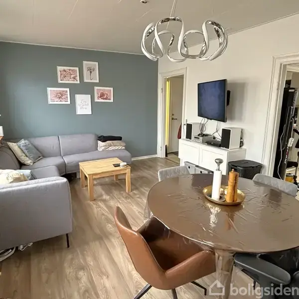 Stue med grå hjørnesofa står mod en blå væg dekoreret med billeder. Rundt spisebord i forgrunden er dækket. TV hænger på væggen ved siden af en åben dør.