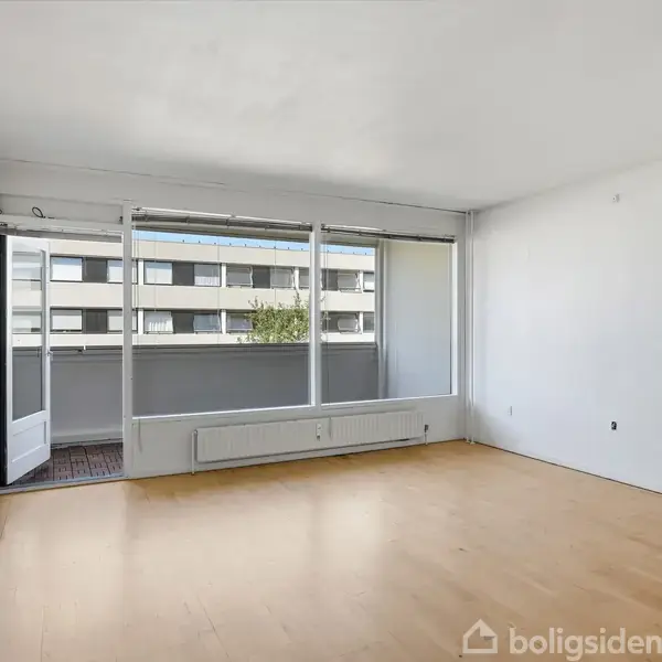 Et tomt rum med trægulv, åbne døre til en balkon. Udsigt over en bygning med mange vinduer gennem store vinduer. Væggene er hvide, og rummet er lyst oplyst.