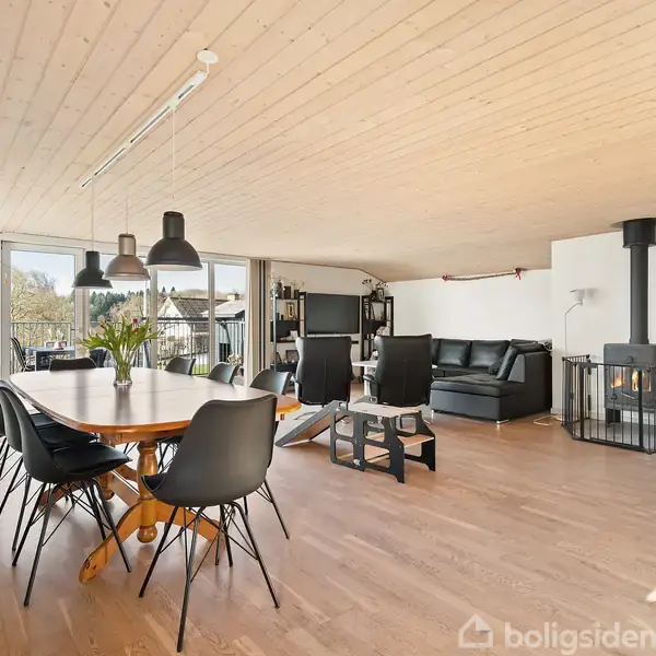Spisebord med sorte stole står i rummeligt opholdsområde med træloft og brændeovn i hjørnet, omgivet af store vinduer og en sort sofa.