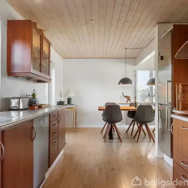 Køkken med træskabe og grå bordplader. Et spisebord med fire grå stole og en lampe hænger over det. Trægulv og lyse trælofter skaber en hyggelig atmosfære.