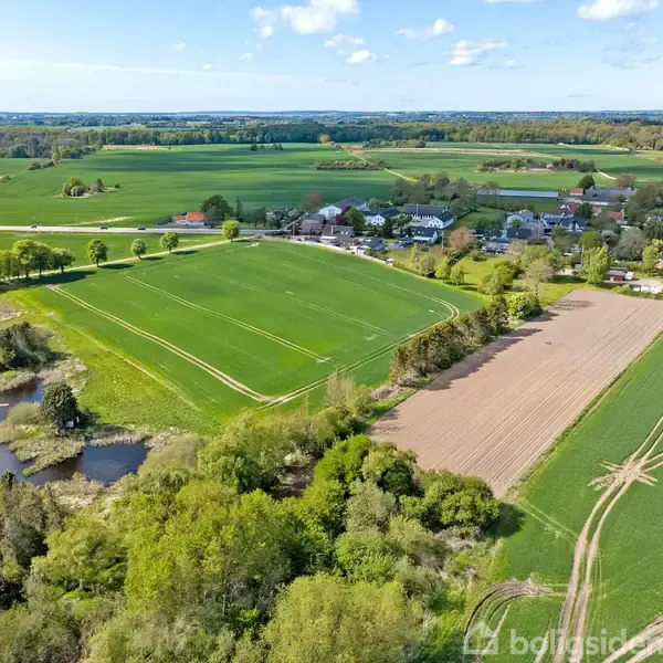 Landskabet viser grønne marker omgivet af træer. En landsby med huse ligger i baggrunden. En lille sø er synlig til venstre, og himlen er klar med få skyer.
