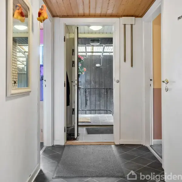 Gang med flisegulv og træloft. En åben dør leder ud til en overdækket terrasse. Spejl på væggen til venstre. Dør til højre markeret "Privat".