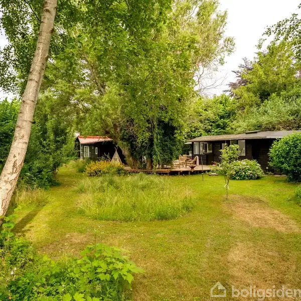 Et træhus står fredeligt blandt frodig vegetation. En træterrasse ved siden af huset giver udsigt over en græsset have med tætte buske og høje træer.