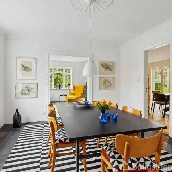 Et moderne spisestue med et sort bord og træstole. En hvid lampe hænger over bordet. Rummet har store vinduer, lyse vægge, og en sort-hvid stribet tæppe.