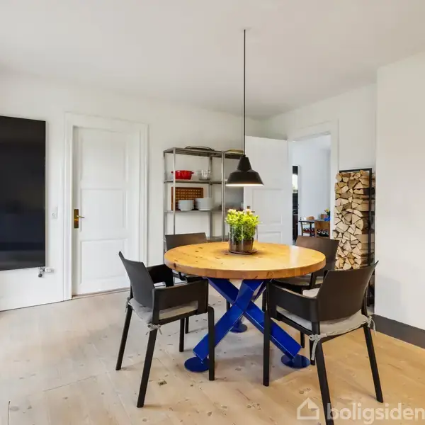 Et rundt træbord med fire sorte stole står i en stue. Bordet har en grøn plante i midten. Til højre ses en pejs med brændende ild.