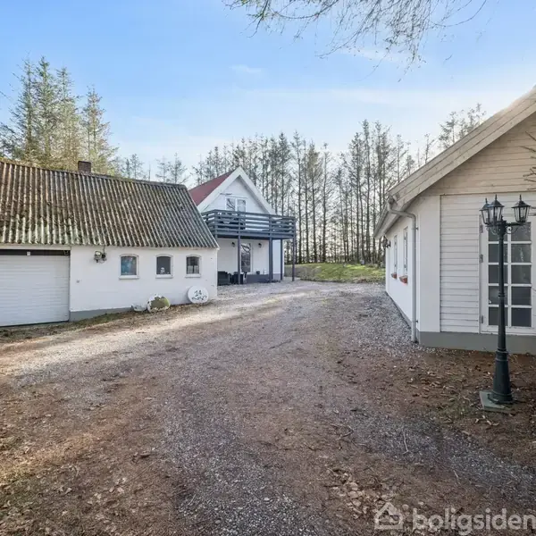 Et hvidt hus med en garage står langs en grusvej, omgivet af træer. I forgrunden ses en lanterne, og i baggrunden er der en stor veranda.