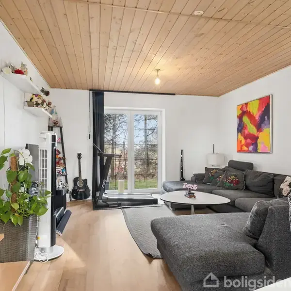 Stue med grå hjørnesofa står i et rum med trægulv og loft. Der er også et sofabord, en guitar, bøger og planter. Store vinduer giver udsigt til en have. Farverig kunst på væggen.