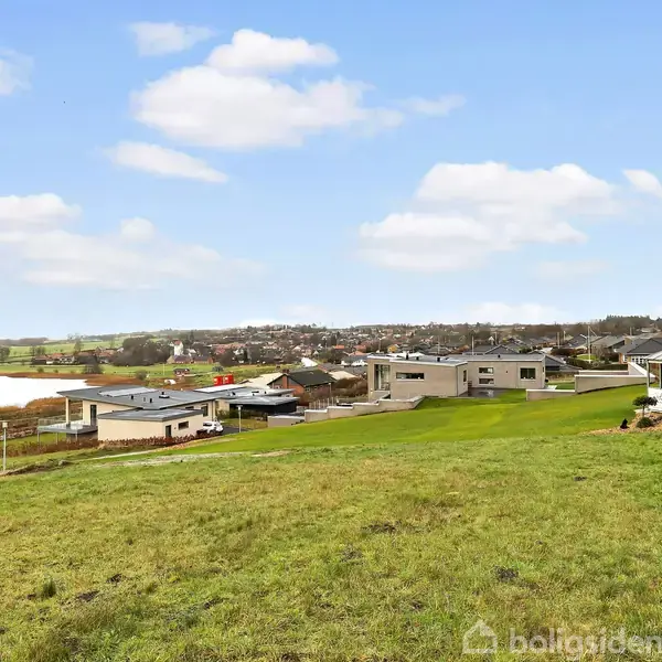 En grøn bakke forrest i billedet vender mod moderne huse i en forstad, med en sø på venstre side og byen i baggrunden under en blå himmel med lette skyer.