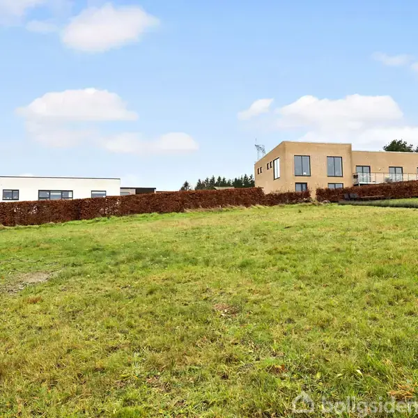 To huse står på en bakke med en grøn græsplæne foran. En hæk adskiller grunden fra resten af området. Himlen er let overskyet med skyer.