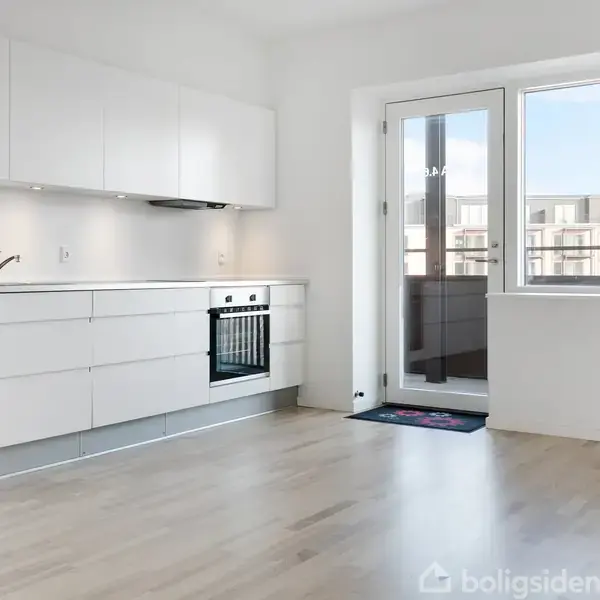 Et moderne, lyst køkken med hvide skabe og indbygget ovn ved siden af en stor, glasdør fører til en altan med udsigt til andre bygninger.