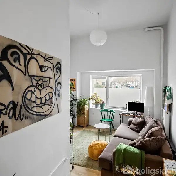 Et maleri af en abe hænger på væggen. En stue med en brun sofa står nær et vindue. Der er grønne planter og stole til pynt.