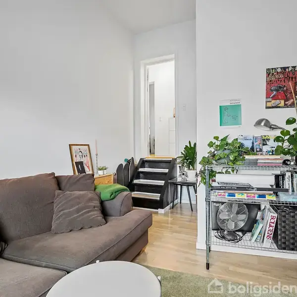 En grå sofa står i en stue ved siden af et hvidt rundt bord. Reolen er fyldt med bøger og planter. En trappe fører til en anden etage.