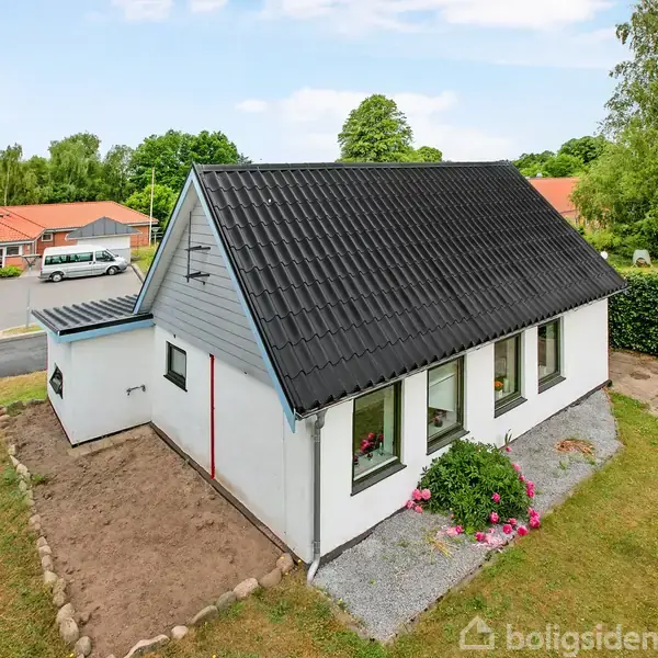 Et hvidt hus med sort tag står omgivet af en have med buske og blomster. I baggrunden ses en lille vej og parkerede biler ved en bygning med rødt tag.
