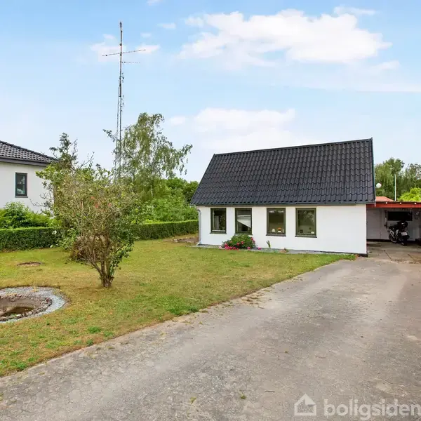 Et hvidt hus med sort tag står stille i en velplejet have. En grusvej fører op til huset, omgivet af grønne hække og en græsplæne med enkelte buske.