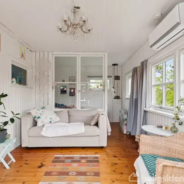 En stue med en lys sofa ved siden af et lille bord med plante; vinduer til højre slipper lys ind, og en kurvestol står foran dem. Tæppe på trægulvet.