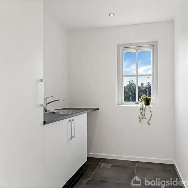 Vask med indbygget skab, står ved et vindue med udsigt til træer. En plante hænger i vindueskarmen. Rummet er hvidt med mørkt flisegulv.