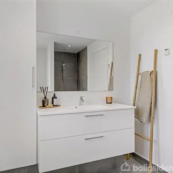 Et moderne badeværelse med en hvid vaskenhed og spejl. Ved siden af er en stige med et håndklæde. Baggrunden har en bruser og vægfliser. En dør ses til højre.