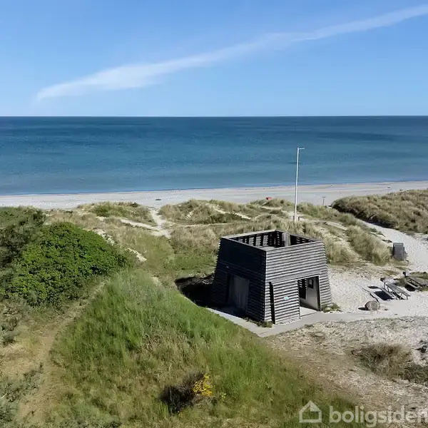 En lille, firkantet, træbygning står stationært blandt sandklitter og græs, nær en bred, sandet strandkant med blåt hav i baggrunden under en klar, blå himmel.