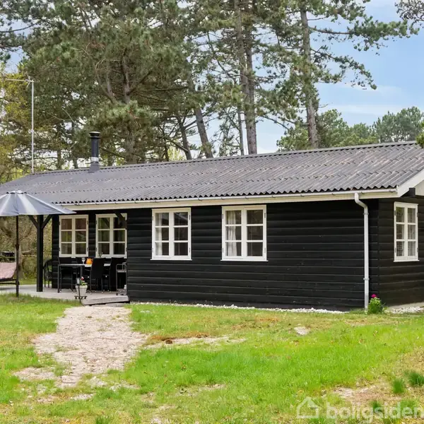 Sort træhus med hvide vinduesrammer står stille i en skovklaring. Husets terrasse har et bord og parasol. Omgivet af græs og træer.