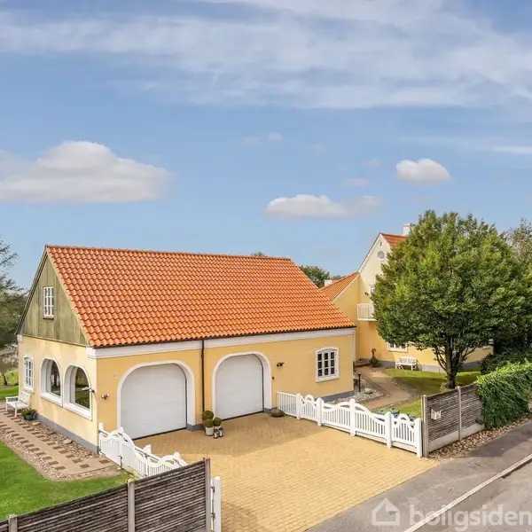 En gul bygning med orange tag står ved vejen, omgivet af grønne træer og en græsplæne. To garageporte ses mod en blå himmel med spredte skyer.