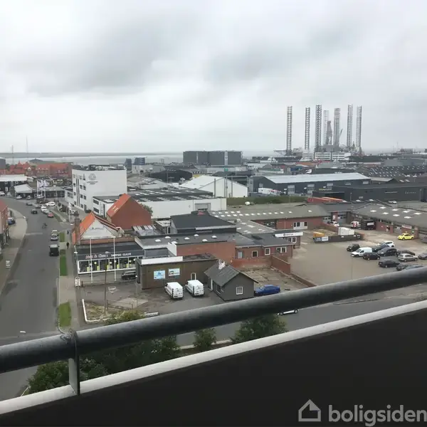 Bygninger i et industriområde er spredt ud omkring en havn med kraner i baggrunden. Gråt vejr og himmel. Udsigt fra en altan med en lav gelænder.