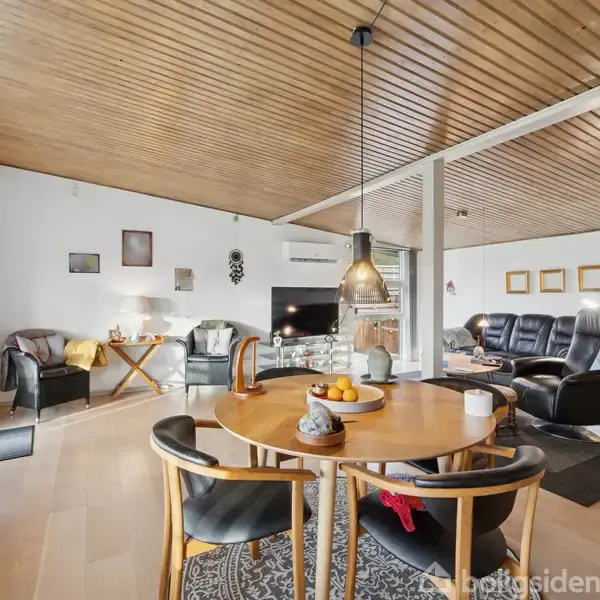 Rundt spisebord med frugtskål i midten af en stue. To sorte stole omkring bordet. Træloft og trægulv. Sofa og lænestole langs væggen. Hyggelig og moderne indretning.