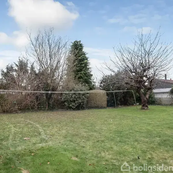 En have med en græsplæne og et tørrestativ i midten. Træer og buske grænser op til haven. Husene ses i baggrunden under en klar blå himmel.