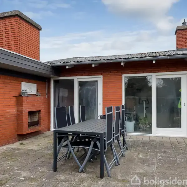 En udendørs terrasse med et sort spisebord og seks stole står foran en rød murstensbygning med to glasdøre. En indbygget grill ses til venstre under en halvtag.