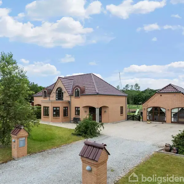 Et rummeligt hus med buet tag og store vinduer står ved siden af en to-bils garage. En grusvej fører til indgangen, omgivet af grønne træer og åben himmel.