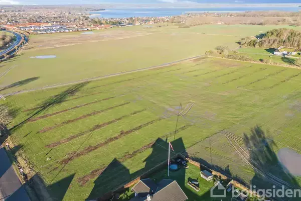 Et stort grønt markområde ligger udstrakt med få bygninger i forgrunden. I baggrunden ses en by mod en skovklædt horisont og en fjord.